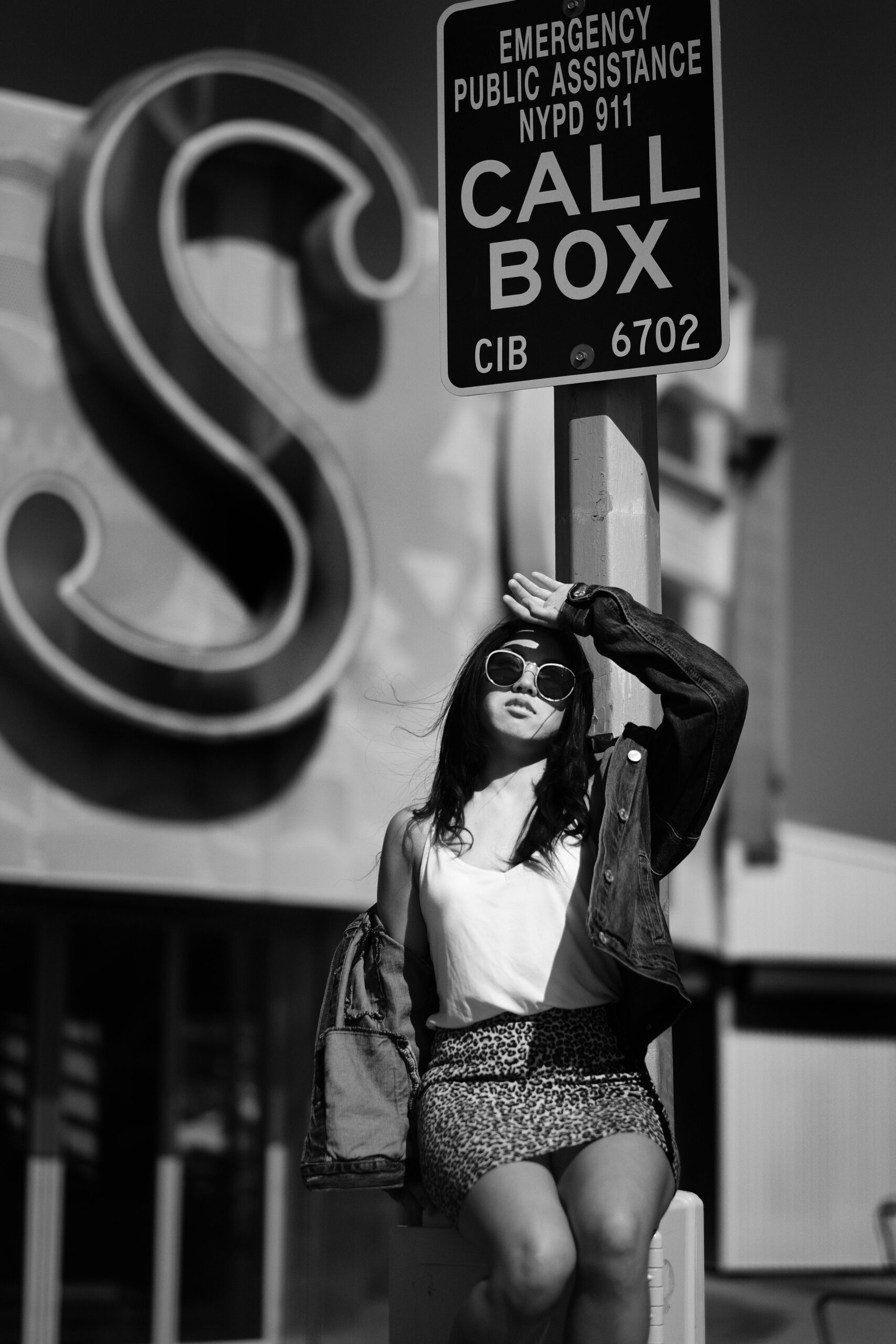 Fashionable woman in sunglasses posing near call box in urban black and white scene.